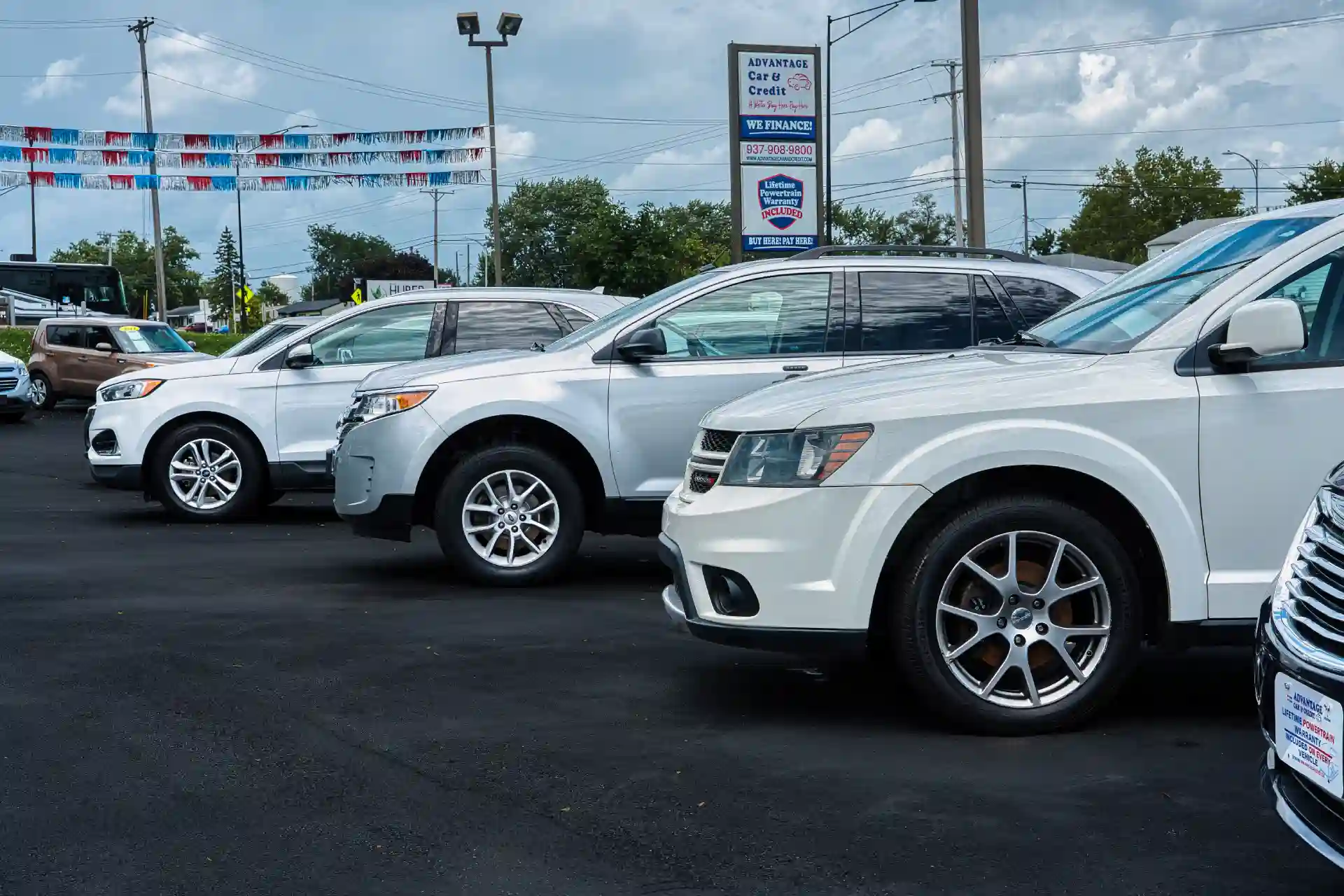 Row of silver and white cars parked at a car dealership lot with a tall sign and decorative pennant banners overhead.